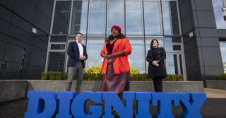 two man and a woman standing behind a blue sign saying dignity- period poverty